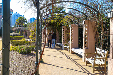 a man and a woman walking through a tunnel of green metal arches covered with lush green plants with light brown stone walls and benches along the path at Atlanta Botanical Garden in Atlanta Georgia