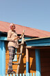 © Stanislaw Mikulski - A girl paints an old metal roof with a roller and a brush while standing on a wooden staircase