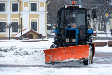 A tractor cleans snow in the city in winter after a snowfall. Cleaning city streets from snow. Tractor driver at work in the city square during a snowstorm.