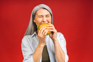 Mature senior woman eating burger with satisfaction. Grandmother enjoys tasty hamburger takeaway, delicious bite of burger, order fastfood delivery while hungry, standing isolated over red background.