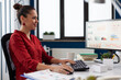© DC Studio - Entrepreneur looking at charts on computer screen in startup office. Smiling businesswoman working on reports at desk. Successful employee in red shirt using desktop pc to analyze business data.