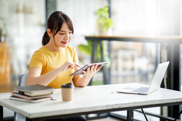 Asian young Businesswoman working and report analyzing financial figures on a graph on a tablet laptop computer sitting in modern coffe shop