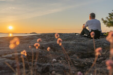 A Man Sits On The Rocks, On The Seashore, He Looks At The Sunset,. The Concept Of Rest, Vacation.