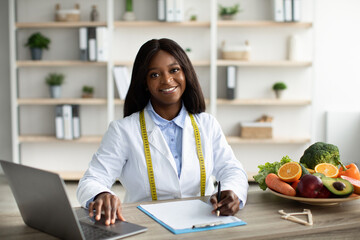 Cheerful african american female dietologist working with laptop, having fresh fruits and vegetables on table