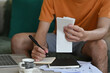 © saltdium - Cropped view of young man in casual outfits sitting on a couch holding receipts and writing down on a paper with a laptop on the side, for business, household expense and technology concept.