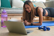 © saltdium - A side view portrait of a young pretty smiling Asian woman planking on a mat in a living room with bottle of water, dumb bells and laptop on the side, for home exercise, online class concept.
