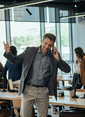 Wall Mural - Happy Businessman Talking on a Mobile Phone in the Office. 
Smiling man speaking on the phone with client while unrecognizable multi-ethnic team of business people working in the background.