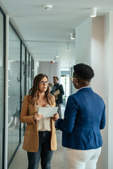 Wall Mural - Serious Businesswoman Holding Digital Tablet and Talking with her Anonymous Female Colleague at Work. 

Two worried business woman standing in modern corridor together and speaking about new project.