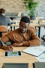 Wall Mural - Handsome African-American businessman analyzing business report and graph while sitting in open plan office table with unrecognizable multi-ethnic team in the background