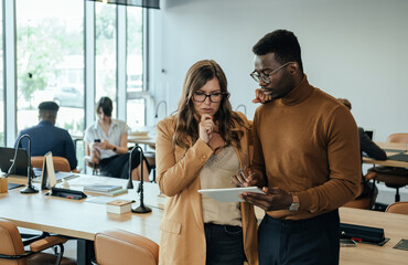 Wall Mural - Two colleagues analyzing business report on a tablet while unrecognizable multi-ethnic team working in the background