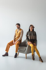 Wall Mural - multiethnic couple in fashionable clothes sitting on armchair and looking at camera on grey