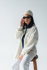 Wall Mural - young asian woman in white jacket and beanie touching dark sunglasses isolated on grey