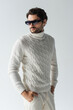 stylish man in white knitted sweater and dark sunglasses standing with hands in pockets isolated on grey
