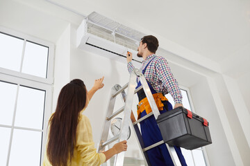 Technician from the AC Maintenance and Repair Service climbs a ladder with his toolbox in order to check or fix troubles with a modern white wall mounted air conditioner in a young lady's home