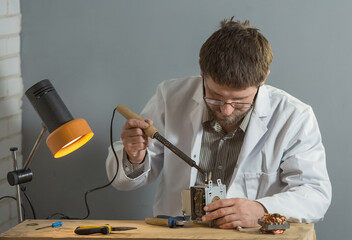 Wall Mural - a man in a white coat is sitting in his hands with a soldering iron and a spare part for an electrical appliance. The concept of assembly and repair of electrical equipment.