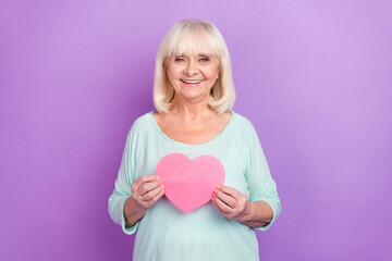 Sticker - Photo of cheerful affectionate old lady hold pink heart shape card wear teal shirt isolated violet color background