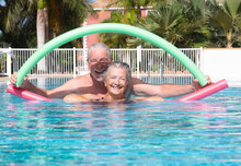 Cheerful Adult Happy Senior Couple Having Fun In Outdoors Swimming Pool Doing Exercise With Swim Noodles. Smiling Retired People Playing Together In The Pool Water Under The Sun