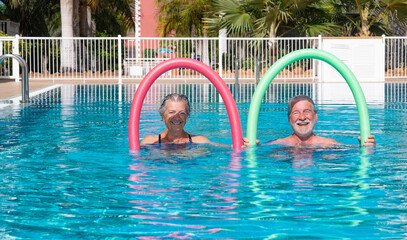 Cheerful adult happy senior couple having fun in outdoors swimming pool doing exercise with swim noodles. Smiling retired people playing together in the pool water under the sun