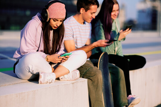 Three teenage friends are sitting in the urban exterior and hanging on their phones.