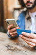 © dzmatph - Soft focus of anonymous male buyer using mobile phone and credit card to pay for online order. while sitting at wooden table in cafeteria in daytime