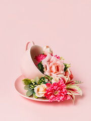 Tea cup and plate with bouquet of fresh roses and orchids on pastel pink background. Creative floral spring bloom concept. Morning drink or healthy breakfast idea. Still life natural visual trend.