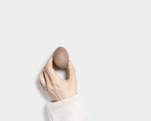 Wall Mural - Symbol Easter beige Easter egg in woman hand. Female arm holding dyed chicken egg brown pastel color, holiday food above white background. Minimal aesthetic photo, Top view