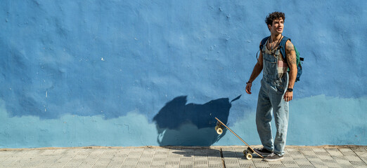 Young handsome skater man with backpack and waveboard. Sunny summer day in the city. Real people lifestyle.