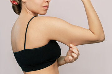 Wall Mural - Cropped shot of a young caucasian woman in a black top grabbing skin on her upper arm with excess fat isolated on a beige background. Pinching the loose and saggy muscles. Overweight concept