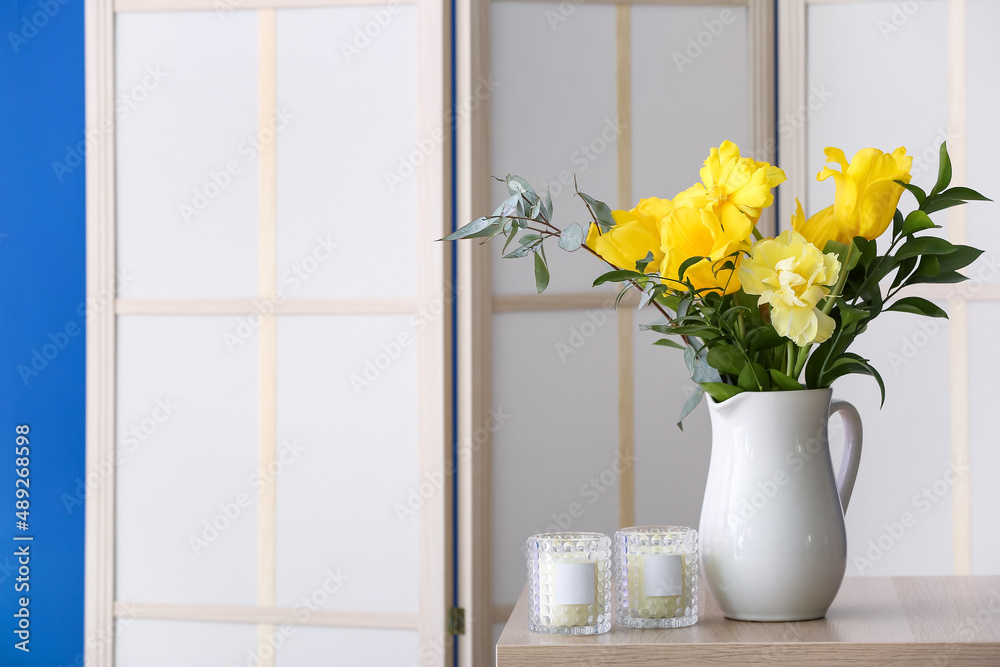 Vase with yellow flowers, candles on table and folding screen near blue wall