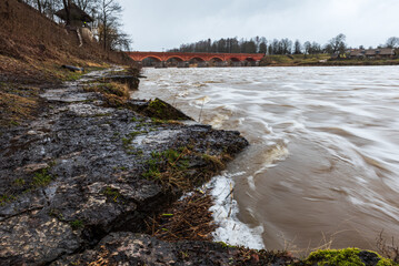 Wall Mural - High water level in Venta river on spring day and old red brick bridge. Kuldiga, Latvia.