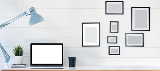 Creative workspace with blank screen laptop computer on white wooden table. Blank picture frame on wooden wall in home office. Mockup home workspace background.