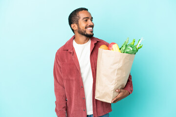 Canvas Print - Young latin man holding a grocery shopping bag isolated on blue background looking side