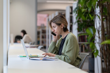 Work from anywhere. Young focused female freelancer talking with client via mobile phone and looking at laptop screen while working remotely in modern cozy library, selective focus. Freelance concept