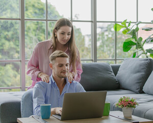 Millennial young Caucasian lover couple handsome husband sitting on floor browsing surfing internet shopping online via laptop computer while beautiful wife on cozy sofa help massaging shoulders.