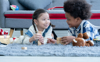 Cute little Caucasian and African kids girl and boy lying on floor smiling and playing toys build wooden blocks together at home. Friendship of diverse ethnicity children