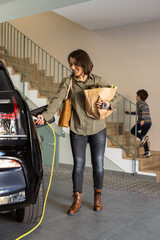 Woman with groceries plugs a charger to electric car socket at home.