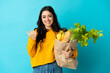 © luismolinero - Young woman holding a grocery shopping bag isolated on blue background pointing to the side to present a product