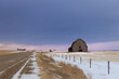 Prairie winter farm road leading into distant grain silos and dark sky. An old barn sits in farm field next to road.