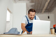 © Kostiantyn - Professional handyman in workwear leaning on hands and smiling while using laptop, standing indoors