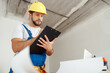 © Kostiantyn - Attentive repairman in workwear and hardhat holding clipboard and making notes while standing indoors during renovation work