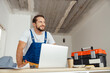© Kostiantyn - Cheerful handyman in workwear leaning on hands and smiling away while using laptop, getting ready for construction work indoors