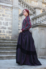 Wall Mural - Full length portrait of red-haired woman wearing a historical victorian gown costume, walking around beautiful location with  Gothic stone architecture.