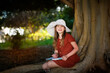 © natalialeb - A cute european kid girl in a dress and a big hat sits near a big tree in the park. A child among the big roots of a tree writes poetry, a girl makes notes