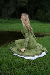 Full length portrait of blondewoman wearing a  beautiful  green medieval fantasy gown. Posing with gestural hands in a enchanted forest background.