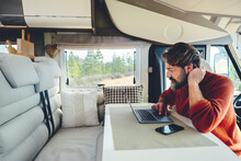 Young Modern Adult Man Work On Laptop Computer In Remote Working And Digital Job Lifestyle Inside A Modern And Connected Camper Van With Nature Woods View Outside The Window. Freedom Lifestyle People