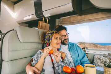 Cheerful mature couple smile and hug sitting inside camper van and enjoy freedom on summer holiday vacation. Ocean view outdoor.. Fruits and healthy food on the table. Tourist people have fun together