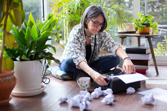 Adult female writer putting new blank sheet of paper on typewriter next to many crumpled papers. Creative block.
