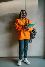 Full Length Portrait Of Smiling Young Woman Holding Books And Backpack Standing Against Gray Wall