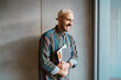 © Maskot - Cheerful male adult student holding book standing in front of gray wall at college