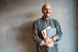 © Maskot - Portrait of cheerful male adult student holding book standing in front of gray wall at college